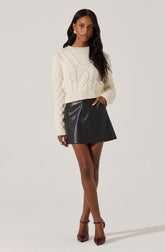 A woman is wearing a cream-colored, chunky knit sweater with a cable pattern and a ribbed hem, paired with a black faux leather mini skirt. She has on brown pointed-toe high heels with ankle straps and accessorizes with gold earrings and rings.